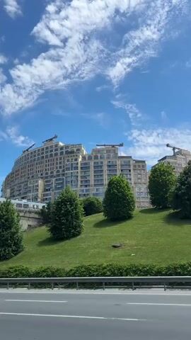 The video shows a ground-level view from a moving vehicle, likely a car or truck, driving on a highway on a sunny day with a blue sky and some clouds. The first shot shows a green highway sign pointing to Gaziosmanpasa, Sultangazi, and Arnavutkoy with modern, large apartment buildings in the background. The camera then moves under a concrete overpass. The final shot shows the side of a blue truck trailer with white lettering that reads, "Bir Yudum Su, Bir Omur Saglik." The background shows the same apartment buildings from the beginning of the video.