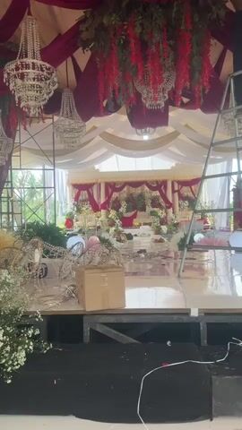 A wedding reception venue is being set up. A man is standing on a ladder, adjusting decorations hanging from the ceiling. The decorations consist of red floral garlands and large chandeliers. Drapes of white and burgundy fabric are hung from the ceiling, creating a canopy. In the background, a decorated stage area is visible with chairs and floral arrangements. The floor is reflective, suggesting it is polished or wet. Several boxes and other setup materials are scattered on the floor in the foreground. The overall scene depicts a work-in-progress for a formal event.