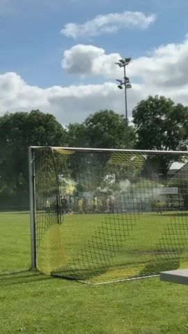 A daytime outdoor shot shows a soccer goal with yellow and black netting in the foreground, partially obscuring the view of a soccer field beyond. Green grass covers the field, and in the distance, a group of people is visible, some of whom are standing amidst a cloud of light green smoke, possibly from a smoke bomb or flare. Tall green trees line the far edge of the field, and above them, a clear blue sky with fluffy white clouds is visible. To the right, a set of stadium lights on poles extends upwards against the sky.