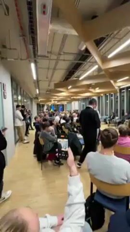 Here is a description of the video:

The video shows a room filled with people seated in chairs. A man in a suit is speaking to a panel of individuals seated in chairs in the front of the room. There are flags in the background. Some attendees are standing around the perimeter of the room. One individual is holding a smartphone, presumably recording the event. The room has a modern design with wooden beams on the ceiling. The floor is a light-colored wood. There are large windows on one side of the room.