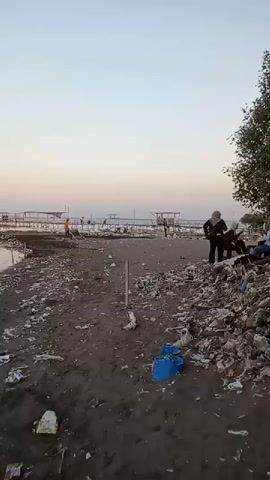 The video shows a shoreline covered in debris, likely plastic waste, with some people present. The sky is a pale gradient of blue and pink, suggesting either sunrise or sunset. In the distance, there are piers or jetties with structures on them, and a few boats or ships are visible on the water. Some individuals are standing or sitting on the beach, while others are near the piers. The overall impression is one of a polluted coastal area with some human activity.