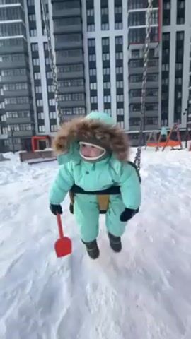 A baby in a light blue snowsuit with a fur-lined hood swings on a playground swing. The baby is wearing a gray balaclava and black mittens, and holds a small red shovel in the left hand. The playground is covered in snow. Behind the swing is a tall, modern apartment building with many windows. In the background are other playground structures and benches, also covered in snow. The camera angle is slightly low, looking up at the baby as it swings back and forth.