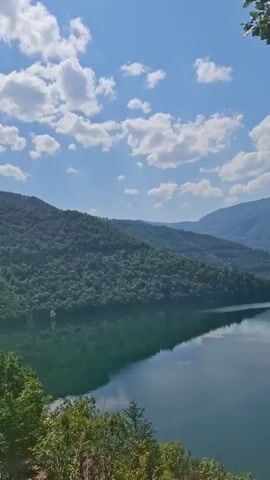 The video opens with a scenic view of a large body of water surrounded by lush green mountains. The sky is a vibrant blue with scattered white clouds. As the camera pans, the water reflects the sky and mountains, creating a serene and picturesque landscape. In the foreground, leafy branches of trees frame the view. The camera then slowly zooms out, revealing more of the winding shoreline and the distant mountain ranges. Towards the end of the video, a woman is seen sitting near a tree, looking out at the view, and holding something to her mouth. The overall scene is peaceful and natural.