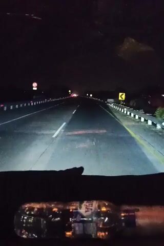 A first-person perspective video shows a nighttime drive on a highway. The camera is mounted inside a vehicle, looking out through the windshield. The road ahead is illuminated by the vehicle's headlights, revealing two lanes in each direction, separated by white lines. The sides of the road are lined with white reflective markers and a guardrail. There are a few distant lights from other vehicles. A yellow directional sign is visible on the right side of the road. At the bottom of the frame, a clear plastic water bottle with a blue label is visible on the dashboard. The bottle reflects the interior lights of the vehicle.
