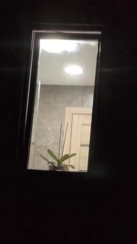 A video shows a dimly lit room through a window. A potted plant with green leaves is visible in the foreground, with a light fixture and tiled walls in the background. A woman with blonde hair tied back enters the frame, turning her head as she moves. The window frame is dark and appears to be part of a door or wall.