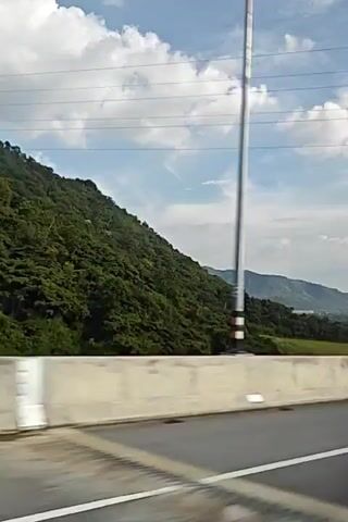 The video shows a highway scene with a mountainous landscape and a cloudy sky. A concrete barrier runs along the side of the road, separating the highway from the vegetation and distant buildings. Several vehicles are visible, including a silver SUV and a pickup truck. The camera appears to be mounted inside a moving vehicle, likely a car, as indicated by the window frame and reflections visible at times. The overall impression is a drive through a scenic area on a clear day.