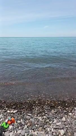A video shows a wide shot of a rocky beach and the sea on a sunny day. A small wave washes up on the shore. Then, two boys appear in the frame. The older boy, wearing gray swimming trunks, bends down at the water's edge, seemingly playing with the pebbles. The younger boy, wearing a white hat and light blue swimming trunks, stands in the water near the older boy. The camera pans slightly to the left, revealing a green, tree-covered cliff in the distance. The sea appears calm with light blue water, and the sky is bright blue with a few scattered clouds.