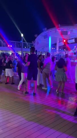 A group of people are dancing on a wooden deck at night. The scene is illuminated by colorful stage lights, including red and blue beams. A band is visible on a stage in the background. Some people are dancing together, while others are dancing individually. The overall atmosphere appears festive.