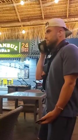 A man in a light-colored baseball cap, gray short-sleeved button shirt, and gray shorts is standing outside a restaurant. He is holding a phone in his left hand and holding a JBL speaker up to his right ear, as if it is a telephone. He smiles, scrunches his face, and speaks into the speaker. He is wearing a crossbody bag. The restaurant has a sign that reads "ISTANBUL KEBAB 24 HOURS OPEN." There are tables and chairs visible outside the restaurant. A young boy in a red shirt and gray pants walks behind the man. The scene is lit by string lights hanging from a thatched roof.