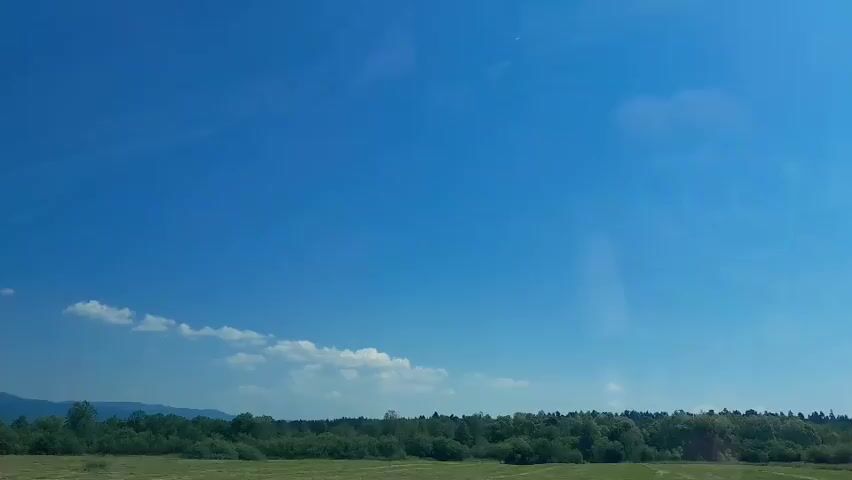The video shows a bright blue sky with a few scattered white clouds. Below the sky is a line of green trees and vegetation. The perspective suggests the video was filmed from a moving vehicle.