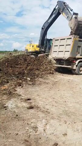 A yellow excavator with a black boom is positioned behind a pile of dirt and debris. The excavator's bucket is extended and positioned over the bed of a large, gray dump truck. The dump truck is facing away from the excavator, with its tailgate down. The sky is blue with scattered clouds. The ground is dirt and appears to be a construction site or outdoor area. The excavator is loading the dump truck with dirt and debris.