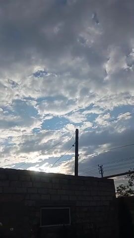 The video captures a cloudy sky over a brick structure and some trees. The sky is filled with a mix of white and gray clouds, with patches of blue sky visible in between. Sunlight is breaking through some of the clouds, creating a dramatic effect. In the foreground, there is a brick wall with a dark rectangular opening that might be a window. A wooden pole with wires extends upwards from behind the wall, suggesting the presence of utility lines. To the right of the pole, another smaller utility pole with multiple wires is visible. Further to the right, the tops of trees are seen, one appearing bush-like and the other a tall, slender evergreen. The overall scene suggests an outdoor setting, possibly a backyard or a rural area, during either sunrise or sunset due to the lighting conditions. The clouds and diffused sunlight evoke a sense of a dynamic or transitional weather moment. There is no visible text in the video.