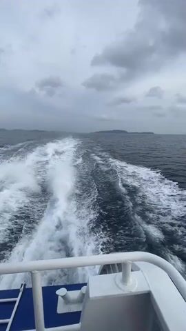 The video shows the view from the back of a moving boat on the ocean. The boat's wake is visible, churning up white foam against the dark water. In the distance, a landmass is visible on the horizon. The sky is overcast with gray clouds. The boat's white railing is visible in the foreground. The camera pans slowly to the right, revealing more of the open ocean. As the camera pans, the sky gradually lightens.