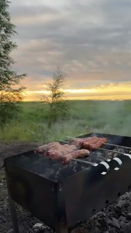The video shows sausages cooking on skewers over a charcoal grill. The grill is in a grassy outdoor setting, with trees and a field in the background. The sky is cloudy with a sunset visible. Smoke rises from the grill as the sausages cook. The camera is positioned at a low angle, looking up at the grill and sausages. The overall mood is relaxed and summery.