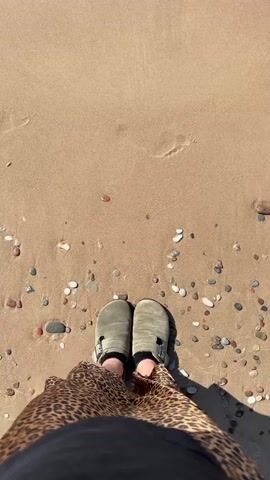 The video shows a pair of feet wearing slip-on shoes and a leopard print skirt standing on a sandy beach. The camera is positioned above the feet, looking down. The tide is coming in, with waves gently washing over the sand and around the feet. Small pebbles are scattered on the beach. The lighting suggests it is daytime.