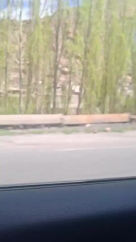 The video shows a view from a car window as it drives past houses and trees in a mountainous region. The houses are simple, stone structures built into the side of the mountain. The trees are mostly bare, with some new leaves emerging. The sky is partly cloudy.