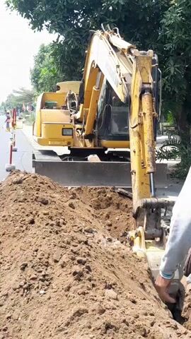 An excavator is digging a trench on the side of a road. The excavator is yellow and has tracks. The trench is filled with dirt. There are trees in the background.