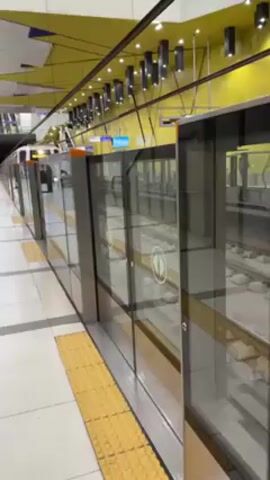 A subway train pulls into a station, stopping at the platform doors which then open. The station platform is tiled, with a yellow tactile paving strip running alongside the platform edge. The platform doors are made of glass and metal. The train is white with orange accents. The station has a yellow ceiling with black light fixtures. A few people are waiting on the platform.