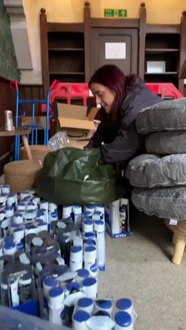 A woman with dark red hair, wearing a dark jacket, is kneeling and reaching into a large green bag. In front of her, there are stacks of Nivea branded products, mostly in spray cans and small jars, many still in their packaging. The products are arranged in rows and appear to be organized for distribution or storage. Behind the woman are wooden shelves and a wooden cabinet with a sign that says "fire exit". The environment appears to be indoors, possibly a storage area or a temporary setup.