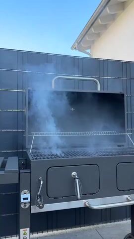 A close-up, low-angle, outdoor shot shows a dark gray grill with its lid open. Thick white smoke billows out of the grill and drifts upwards. The grill has multiple grates inside, and there are visible flames or glowing embers beneath the grates. A silver handle is attached to the front of the grill, and another silver handle is on the side. The grill is positioned in front of a dark gray, slatted fence. Above the fence, a bright blue sky is visible, with the edge of a white, sloped roofline in the upper right corner. The scene is static, with no visible people or animals.