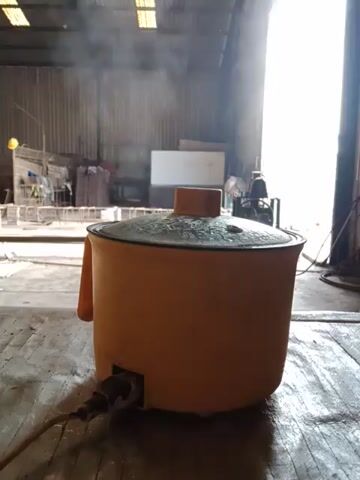 The video shows a yellow cylindrical container with a glass lid on a wooden floor. A power cord extends from the container. Steam is rising from the top of the container. The background features a large warehouse-like space with corrugated metal walls and a forklift or other vehicle parked further back. Sunlight is visible through an opening at the end of the building.