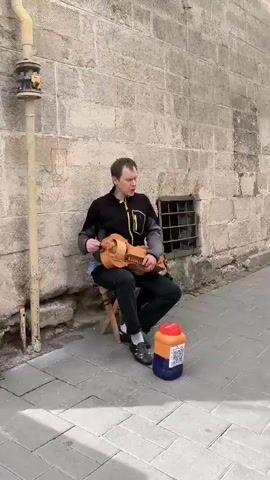 A man sits on a stool playing a hurdy-gurdy against a stone wall. He is wearing a black and grey jacket with a yellow zipper and black pants. Next to him is an orange and blue plastic container with a white label. The wall has a metal grate over a small window. The ground is paved with grey and white tiles. A metal pipe runs vertically up the left side of the frame.
