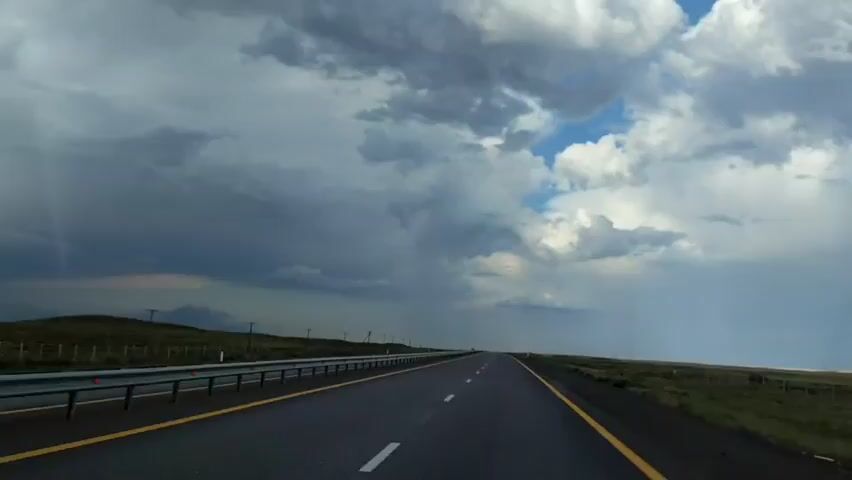 This is a first-person perspective video taken from inside a vehicle traveling on an empty highway. The road has two lanes in each direction, separated by a median with a guardrail. The sky is dramatic, with dark, heavy clouds dominating the left side of the frame, suggesting an approaching storm. To the right, there are patches of blue sky and lighter clouds, with some areas appearing to have rain falling. The landscape on either side of the highway is relatively flat and appears to be grassland or open fields, with some sparse vegetation and a fence line visible. In the distance, there are faint outlines of hills or mountains. The sun is not directly visible, but its light illuminates the scene, casting shadows and highlighting the texture of the clouds. There are no other vehicles or people visible in the immediate vicinity, creating a sense of isolation and vastness. The overall atmosphere is one of impending weather change.