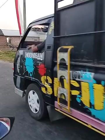 A black pickup truck with a metal cargo bed is driving down a paved road. The truck has colorful graffiti on its sides, including the word "SFUTURALB3." The truck's license plate is visible and reads "AG. 8966 DB." The road is lined with grass and some trees on the left side. On the right side, there are houses and a flag. The sky is overcast. The video is filmed from the perspective of someone on a motorcycle, which is partially visible at the bottom of the frame.