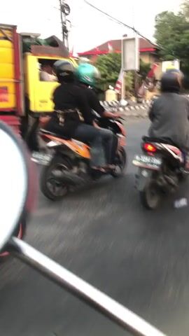 In a medium shot captured from a slightly low angle, two individuals are riding a motorcycle. The person in the front wears a black helmet and a black long-sleeved shirt over blue jeans. The person in the back wears a light blue helmet and a black long-sleeved shirt, also over blue jeans. They are seated side-by-side on an orange and black motorcycle. The motorcycle has a license plate with the text "AD 6023 AGU".

To the left of the motorcycle, a yellow truck with red trim is partially visible. The truck is moving. The scene appears to be an intersection with streetlights and traffic signals. In the background, there are buildings, some with red roofs, and trees. A sign with Indonesian text is visible, reading "POLSEK CANDI". The lighting suggests it is either early morning or late afternoon, with soft, diffused light. The overall atmosphere is that of everyday traffic in a semi-urban or rural area in Klurak, Indonesia. The movement of the vehicles and the presence of traffic infrastructure indicate an active road. No specific emotions are discernible from the subjects due to the distance and resolution.