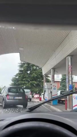 The video is filmed from inside a car, looking through the windshield. The car is at a gas station, waiting in line. There are other cars in front and to the side. A large pine tree is visible to the left, and gas pumps are to the right under a canopy. The ground appears wet, suggesting recent rain. The sky is overcast.