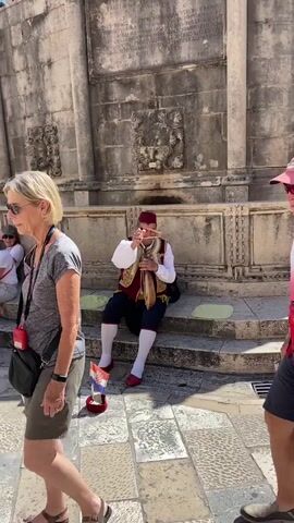 The video shows a street performer in traditional attire playing a musical instrument while tourists walk by. The performer is sitting on steps in front of a stone building, wearing a red fez, a vest, and a white shirt. He is holding a wooden instrument with strings. In the foreground, two tourists are walking past, one is wearing sunglasses and a red and white shirt, and the other is partially visible on the left. A woman is sitting on the steps in the background, observing the performer. The environment appears to be a sunny, outdoor location with stone architecture.