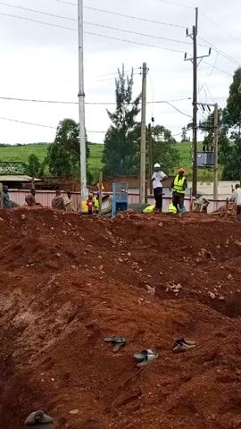 The video shows a construction site with several people working. The site is filled with reddish-brown soil, with a deep trench visible.  Some workers are wearing helmets and safety vests. Some are digging and moving material in yellow containers. There's a pile of gravel, and a building in the background with a sign that reads "THE SALUTE PLACE".  The sky is overcast. The camera moves a little, and several women in casual clothing are walking on the hill, one woman is wearing a blue headscarf and looking directly at the camera.  Boots and sandals are strewn on the ground.  Power lines and poles are visible.