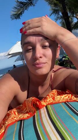 A woman is lying on a beach towel at a beach resort. She is wearing a maroon bikini, has red nail polish, and a bracelet on her right wrist. She has her eyes closed and her right hand on her forehead, partially obscuring her face. The beach towel is brightly colored with orange, yellow, blue and green stripes and patterns. Behind her, there are beach umbrellas, palm trees, and a blue ocean. The sky is clear and sunny. The camera appears to be held by the woman, capturing a selfie-style view.