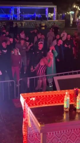The video shows a nighttime outdoor event with a crowd of people. In the foreground, there is a table covered with a red and white patterned cloth. Two bottles are visible on the table. A metal barrier separates the table from the crowd. A person in a black and pink striped singlet and shorts is dancing in front of the crowd. Other people in the crowd are watching, some are holding up phones. In the background, there is a stage with lights and a banner that appears to say "FEST". The environment appears to be a festival or an outdoor concert.
