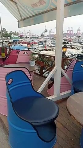 This video pans across an outdoor seating area overlooking a harbor. The seating area features a wooden deck with round wooden tables and chairs made from repurposed blue and pink barrels. Potted plants and lamps are placed on the tables. In the background, numerous boats are docked in the harbor, and large industrial cranes are visible on the shore. The sky is overcast, suggesting a cloudy day. The camera movement is slow and steady, providing a panoramic view of the setting.