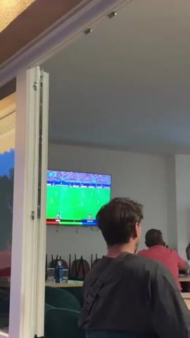 The video shows a group of people watching a soccer match on a large television screen inside a room. The screen displays a soccer field with players in red and white uniforms. In the foreground, a young boy wearing a white soccer jersey with the number 5 on the back is standing and watching the game with enthusiasm. He is looking up at the screen and smiling. Behind the boy, a man with dark hair is sitting and also watching the game. Other people are visible in the background, and there are chairs stacked against the wall. The room appears to be a restaurant or bar, with tables and drinks on them. The overall atmosphere seems relaxed and engaged, with people enjoying the sporting event.