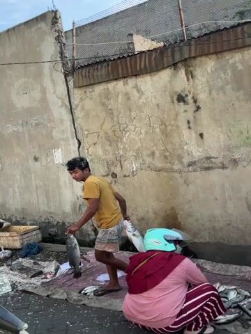 A man in a yellow t-shirt and shorts carries two large fish, one in each hand. He stands in front of a weathered concrete wall. To his right, a person wearing a white shirt and a blue cap sits on a motorbike, facing away from the camera. The ground is littered with debris and some fish. A second person, partially visible in the lower right of the frame, is also seated on the motorbike. The scene appears to be an outdoor market or roadside area.