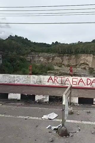 The video shows a view from a moving vehicle. The scene depicts a roadside barrier with graffiti on it, a riverbed with rocks and debris, and a hillside with trees and eroded land. The graffiti on the barrier includes the words "ARTAGADA", "3117-01", a heart symbol, "22 GARD", "ZENI KONSTRUKSI", and "THE BEE SOLDIER". The sky is overcast, suggesting a cloudy day. The overall impression is one of a somewhat neglected or damaged environment.