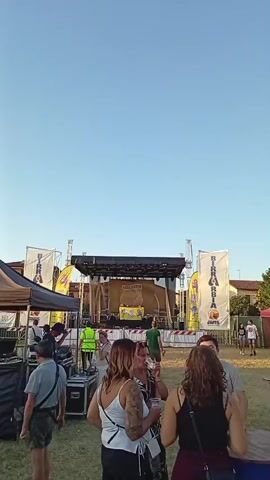 This is a vertical, outdoor video, filmed at dusk. The sky is clear and blue. In the background, a large stage is set up with a banner that reads "DIRACCHIA JONIS." There are several people standing around the stage, and equipment is visible, including what appears to be DJ decks. To the left of the stage is a brown tent with black drapes. In the foreground, several people are gathered, some are talking and holding drinks. Further in the background, to the right, an inflatable playground slide is visible, with people milling around it. The ground is covered in grass, and there are some trees and buildings in the distance. The overall atmosphere appears to be that of an outdoor festival or event.