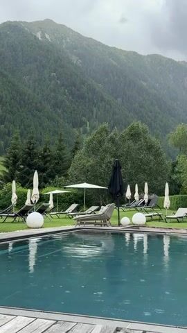 This media file depicts a serene outdoor swimming pool area with a stunning mountain backdrop. The foreground features a clear, dark blue swimming pool, with visible ripples indicating rainfall. The surface of the pool reflects the overcast sky and the surrounding greenery.

Beyond the pool, a manicured lawn is dotted with numerous empty lounge chairs and closed white umbrellas, suggesting a pause in recreational activity due to the weather. A few large, white spherical decorative lights are strategically placed on the lawn. One black umbrella is open, offering a stark contrast to the white ones. The lounge chairs and umbrellas are neatly arranged, indicating an organized and well-maintained space.

In the mid-ground, a dense forest covers the lower slopes of a majestic mountain. The trees are predominantly evergreen, creating a lush, deep green tapestry. The mountain itself is shrouded in mist and appears to be receiving rain, with visible streaks of water descending its face. The sky is heavily clouded, contributing to the muted and somewhat melancholic atmosphere.

The overall setting evokes a sense of tranquility and natural beauty, characteristic of a mountain resort or spa. The rainfall adds a dynamic element to the scene, creating a peaceful, almost meditative ambiance. The absence of people and the closed umbrellas suggest that the moment captured is during a rainy spell, likely in the late afternoon or evening given the overcast conditions and diffused light. The location cues point to Mühlbach - Rio di Pusteria, Italy, a region known for its mountainous landscapes and outdoor recreational facilities. There is no visible text in the media file.