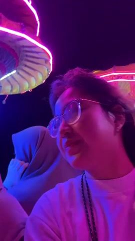 The video begins with a close-up of a young woman wearing glasses, her face illuminated by pink and purple lights. In the background, another woman wearing a headscarf is visible, smiling and waving. The scene appears to be outdoors at night, with colorful lights adorning what looks like a festive structure. The camera then pans to the left, revealing a street scene with buildings and trees. A black SUV is parked on the side of the road. The camera continues to move, showing a blurred street with more lights and people on motorcycles. The video appears to capture a lively night out, possibly at a festival or carnival.