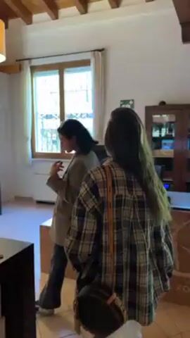 Two women are in a wine shop in a sunny room. One woman wears a beige blazer and black pants. The other wears a plaid overshirt and white pants. They are standing by a counter that has wine bottles on it. Behind them is a wall of wine bottles in shelves. The woman in the blazer is pointing at the bottles and talking to the woman in the plaid shirt.