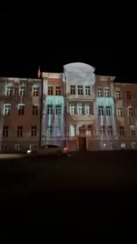At night, a large three-story building with many windows is illuminated with a light show. The building is light-colored with a dark roof, and is set against a completely black background. The light show projects various patterns and shapes onto the building's facade, including lines, colors, and white shapes that give the impression of water flowing. A car with illuminated red tail lights is parked on the left side of the screen. A few people, including one pushing a stroller, are walking along the sidewalk in front of the building.