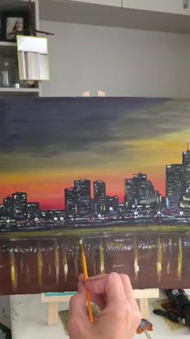 A person's hand is holding a paintbrush and is applying paint to a canvas. The canvas is on an easel and depicts a city skyline at sunset or dusk. The sky is a mix of dark gray, yellow, and orange. The city buildings are silhouetted against the colorful sky, with lights visible in the windows. Below the skyline, there is a reflection of the city lights in the water. The person is wearing a ring on their left ring finger. The background shows shelves with various objects.