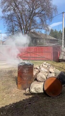 The video shows a rusty metal barrel that is burning contents inside, creating white smoke. The barrel is sitting in a grassy yard next to a pile of rocks. The yard is surrounded by a red wooden fence, and behind the fence is an old wooden house. A large tree stands to the left of the barrel. The sky is blue with wispy clouds. There are no people visible in the video.
