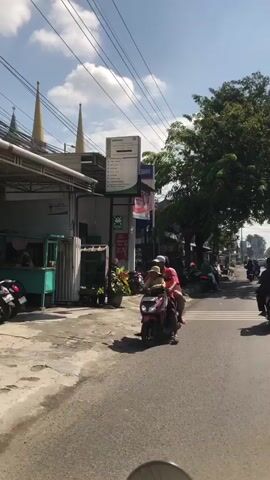 The video shows a street scene in what appears to be a populated area, possibly a town or city in a warm climate. The time of day is daytime, with clear skies and bright sunlight, casting shadows. In the foreground, the bottom of a vehicle, possibly a motorcycle, is visible, suggesting the video is shot from a moving vehicle.

The street is paved and appears to be a two-lane road. On the right side of the street, traffic is moving, with several motorcycles and at least one car visible in the distance.

On the left side of the street, there is a building with a white facade and a green awning. Adjacent to it, there is a sign with text and logos, possibly advertising a business or institution. Further down the sidewalk, there are parked motorcycles and some vegetation. Two minarets or spires are visible above the building.

The main focus of the scene is a motorcycle carrying three people: a driver, a passenger behind them, and a child seated in front of the driver. They are riding on the road, heading away from the camera. The driver and the passenger behind are wearing casual clothing, and the child is wearing a yellow shirt. The motorcycle is red.

Further down the street, there are more buildings, trees, and a general sense of daily life and activity. The environment appears to be somewhat cluttered, with power lines visible overhead.