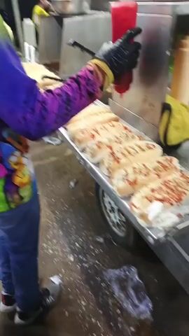 A young man wearing a purple and green Simpson-themed shirt, blue jeans, and a white baseball cap, stands at a food cart preparing hot dogs. He wears black gloves and is using a red squeeze bottle to apply ketchup to the hot dogs lined up on a stainless steel surface. The hot dogs are already topped with shredded cheese and another sauce. Behind him, another person is visible operating a deep fryer. The setting appears to be an outdoor food stall at night. Music is playing in the background.