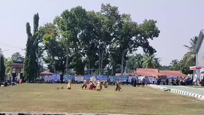 A group of performers in traditional attire are dancing on a grassy field in front of a large building with a tent setup. The building appears to be a school or community center. There are banners displayed behind the performers, and spectators are seated under the tents and standing along the edge of the field. The sky is clear and the sun is shining.