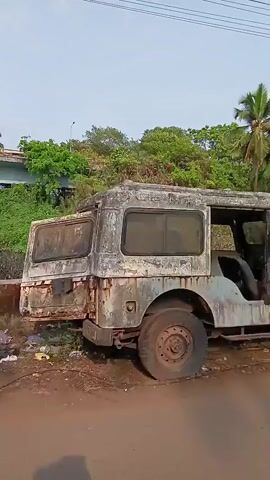 The video shows two derelict vehicles parked on the side of a road. The first vehicle is a heavily rusted, off-road type vehicle with a hard top, missing paint, and a flat tire. The second vehicle is a small hatchback car covered in dirt and overgrown with plants.  The road is unpaved with some debris. Lush green vegetation, including palm trees, fills the background. Overhead power lines are visible against a light blue sky.  The scene appears to be in a tropical or subtropical location. There are no people visible in the video.