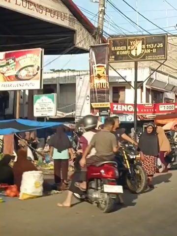 The video shows a busy street scene, likely a market. In the foreground, a man on a motorbike with gold accents is turning, with a passenger behind him. Several other motorbikes are parked or moving. People are walking and interacting. A woman in a hijab and patterned dress is standing near the motorbike. Signs are visible, including one with the word "RAITOR" and another advertising "ZENGA KRETEK" for 7,000 BKS. A black van with a license plate starting with "BL-8227" is also present. Further in the background, there are market stalls with various goods, possibly produce, covered by tarps and umbrellas. Buildings line the street, and there are numerous electrical wires overhead. The overall atmosphere appears to be that of a commercial area with a lot of pedestrian and vehicle activity.